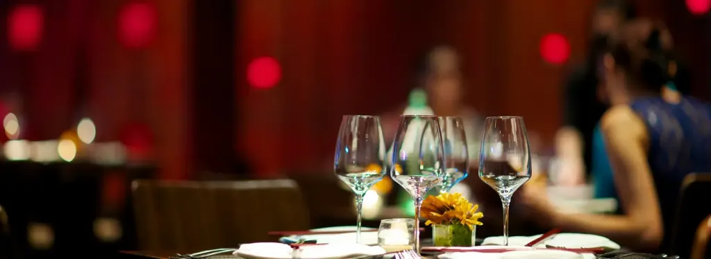 Mesa elegante en Margarita con copas de vino vacías, vela encendida y una flor amarilla sobre mantel blanco, con fondo desenfocado de comensales.