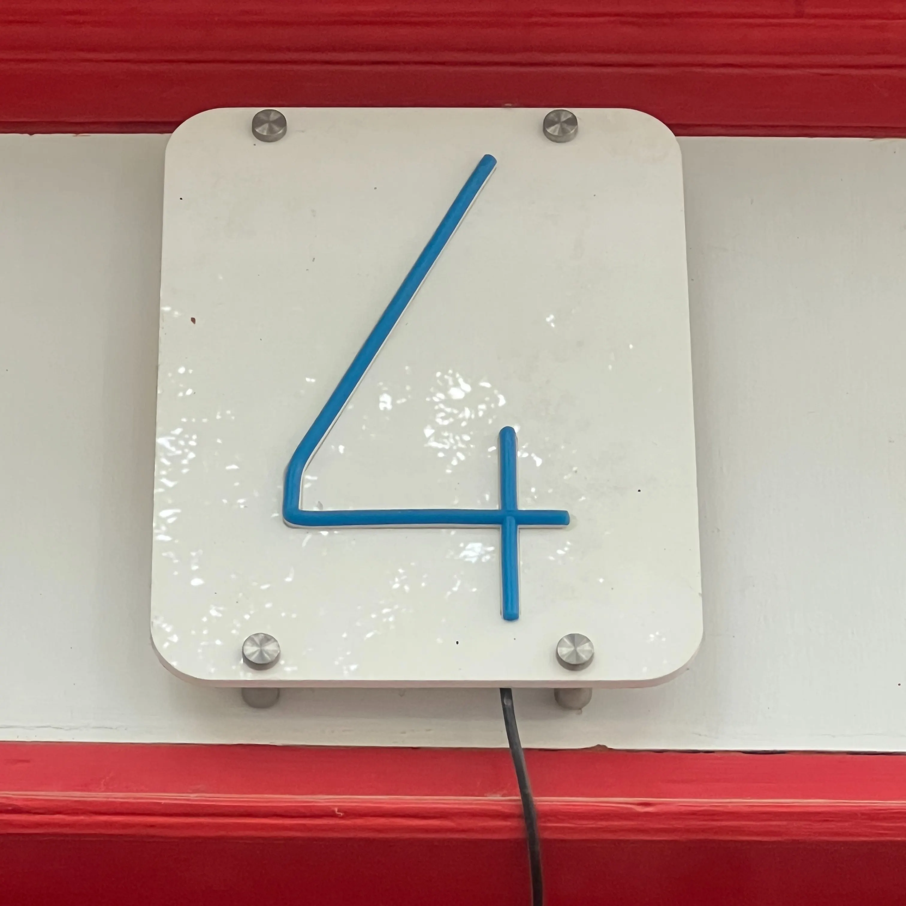 Placa blanca con el número 4 en azul de estilo moderno, fijada con tornillos metálicos y un cable negro visible en la parte inferior.