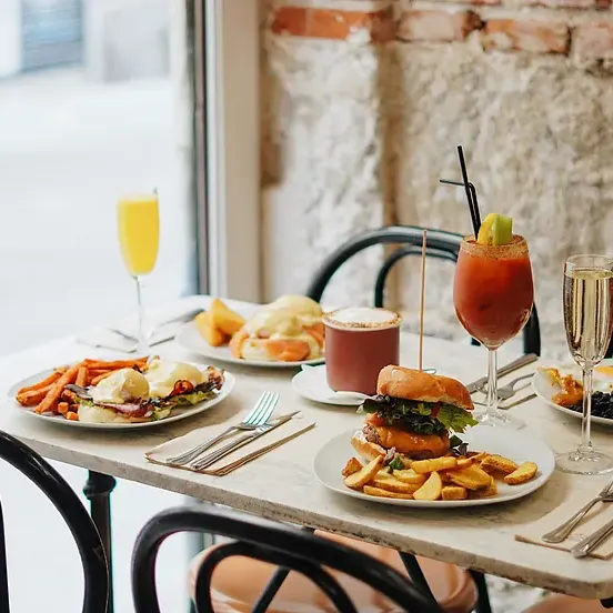 Mesa para cuatro con hamburguesas, papas normales y camote, y cuatro bebidas variadas; fondo con pared de ladrillo visto.