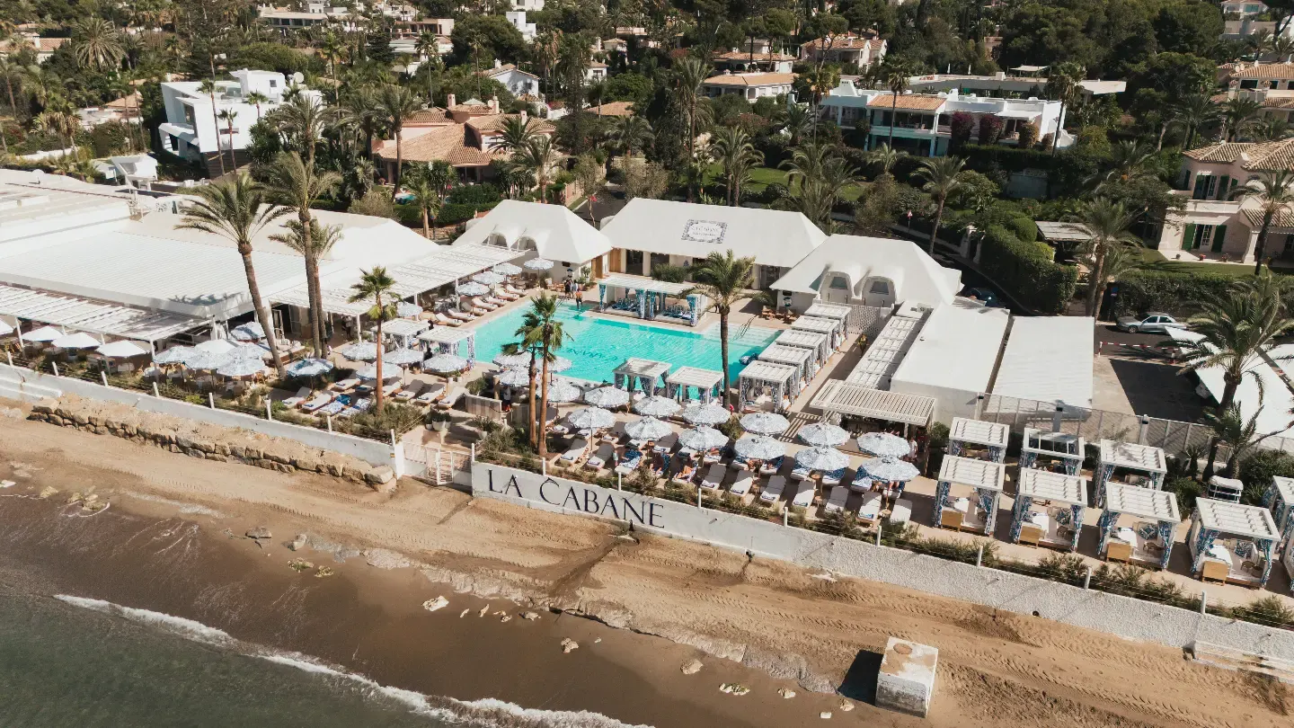 La Cabane Marbella Beach Club destaca por su exclusiva piscina frente al mar, sombrillas elegantes y vistas a la playa rodeada de palmeras.