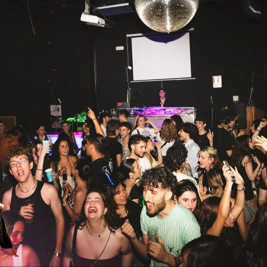 Discoteca llena con gente bailando y socializando. Cabina de DJ al fondo, bola de espejos en el techo y luces de colores sobre la multitud.