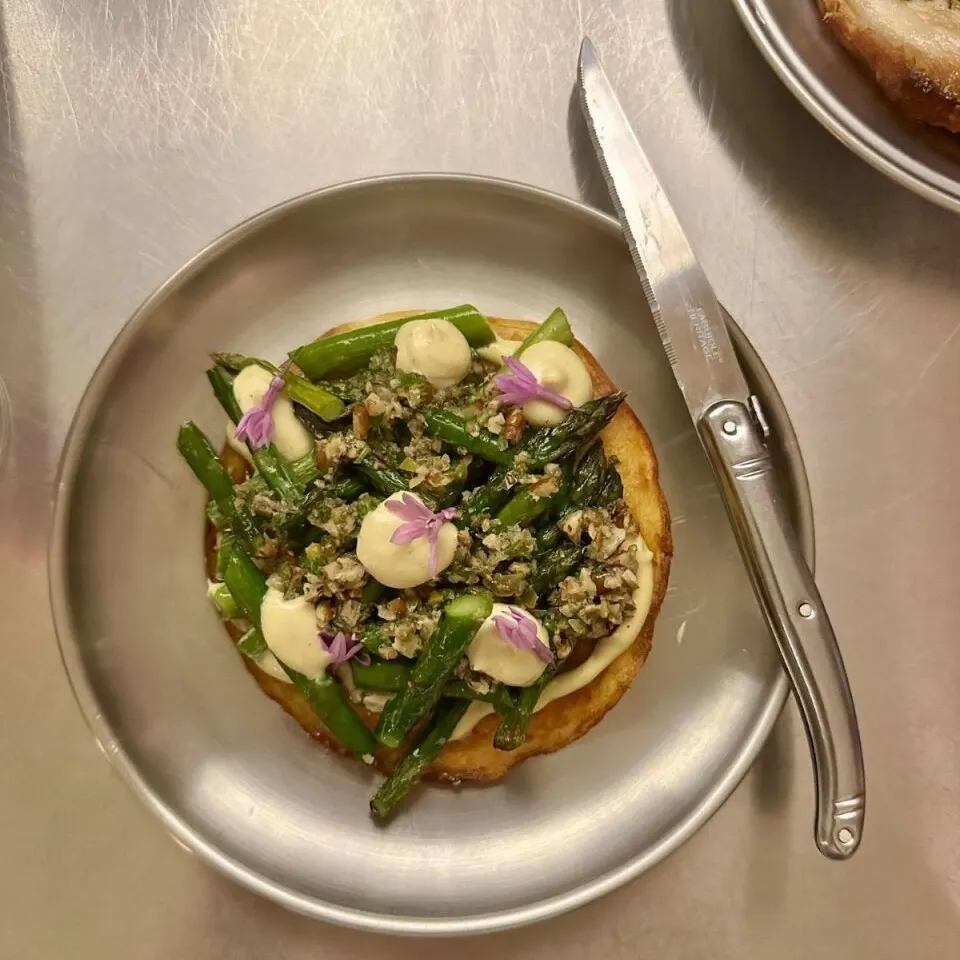 Plato con base redonda, espárragos verdes, crema en pequeños puntos y flores comestibles moradas. Cuchillo a la derecha y otro plato al fondo.