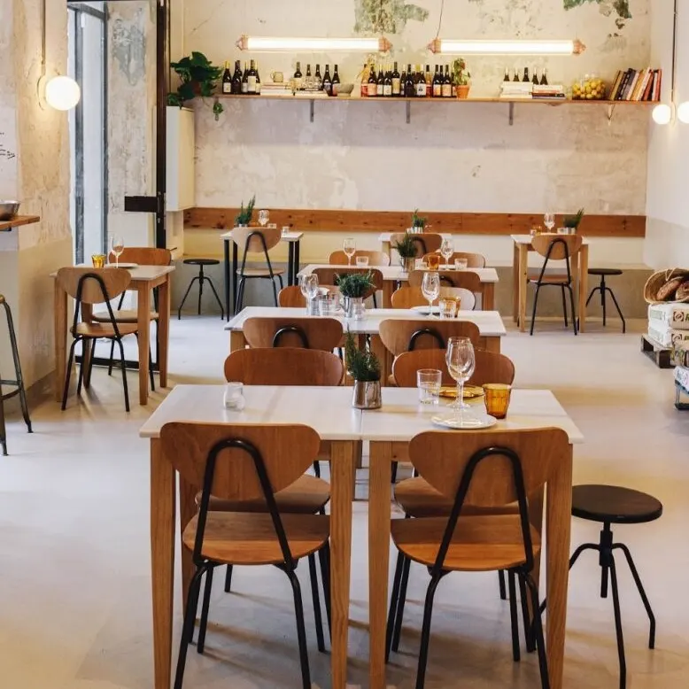 Café minimalista con mesas de madera, plantas, pared rústica de hormigón, estantería decorativa y luces cálidas colgantes.