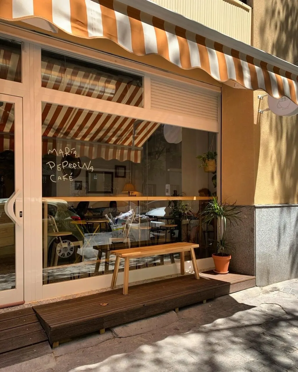 Fachada de María Peperina Café con toldos a rayas naranjas y blancas, banco de madera en la entrada y plantas decorativas junto al ventanal.