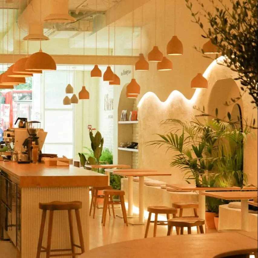 Interior moderno con mesas de madera, plantas, luces colgantes, barra con taburetes y cafetera en ambiente acogedor.