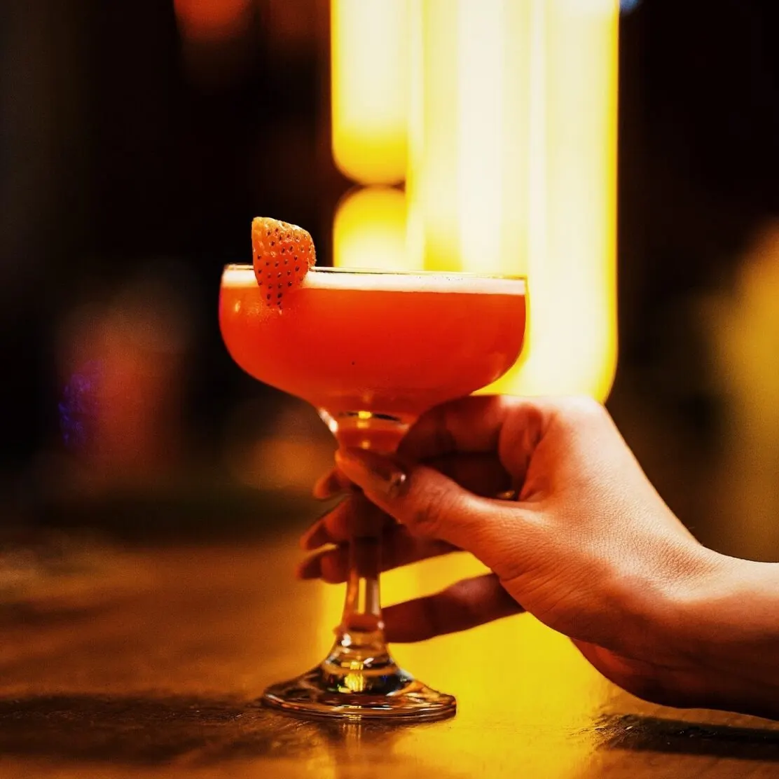 Mano sostiene copa con cóctel rojo anaranjado y fresa en el borde, fondo desenfocado con luces cálidas.