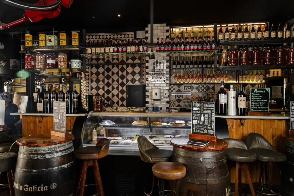 Interior de bar rústico con barriles como mesas, vitrina con comida, estanterías con botellas y decoración con azulejos y pizarras. Ambiente acogedor.
