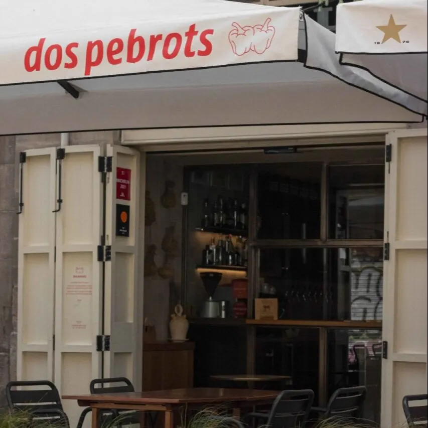 Fachada de dos pebrots con toldo blanco y rojo, puertas abiertas, mesas exteriores y macetas con plantas verdes.