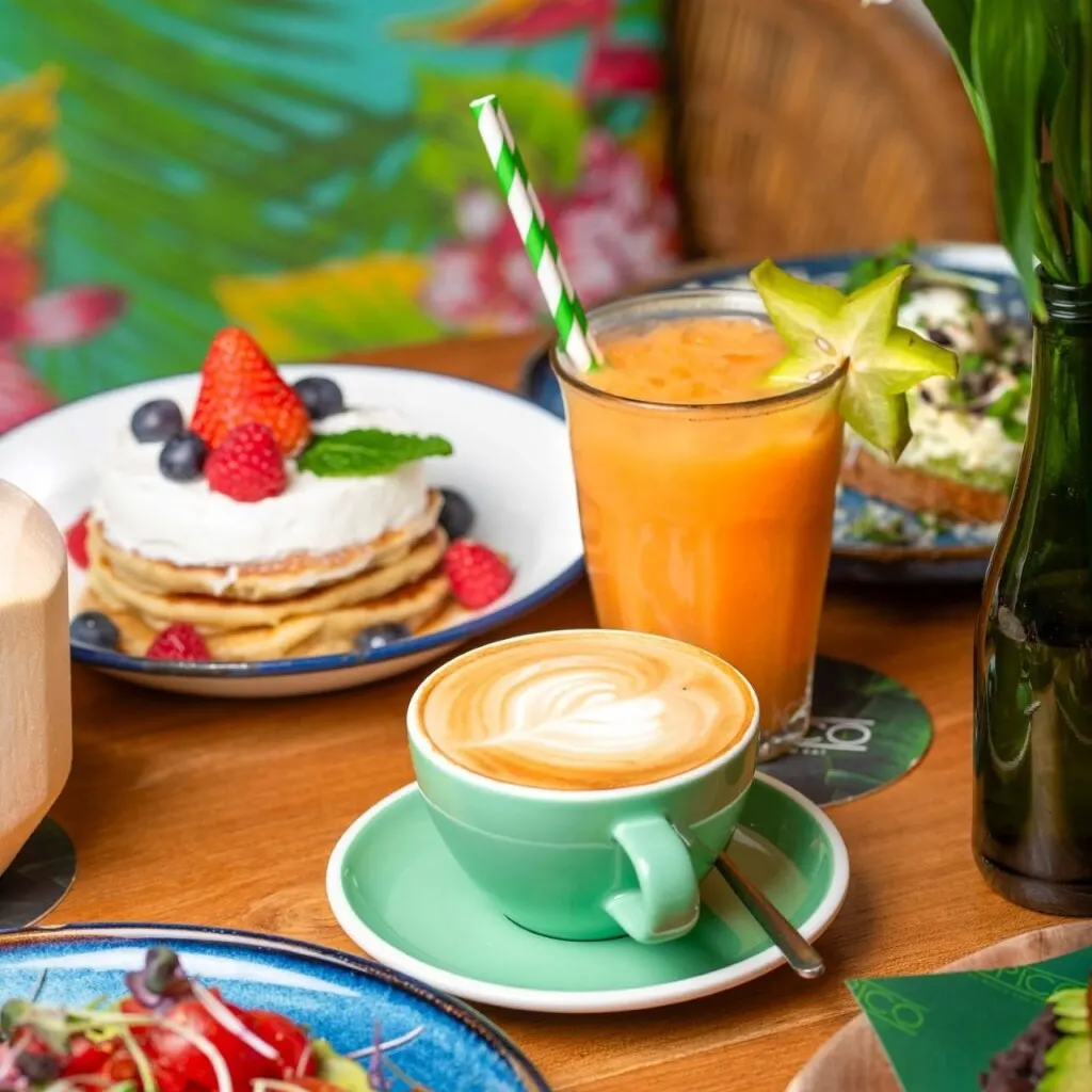 Pancakes con nata, frutos rojos y menta, zumo con carambola y cappuccino con arte latte en café de decoración tropical.