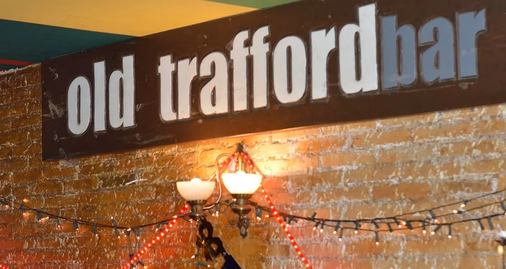Cartel de madera con el nombre de old trafford bar sobre una pared de ladrillo visto, decorada con luces de colores y una lámpara vintage de pared.