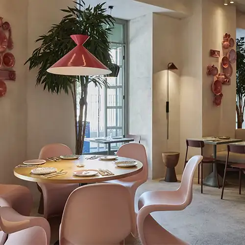 Interior de restaurante con decoración colorida, mesas preparadas, techos ondulados con luces LED y barra con botellas y vegetación colgante.