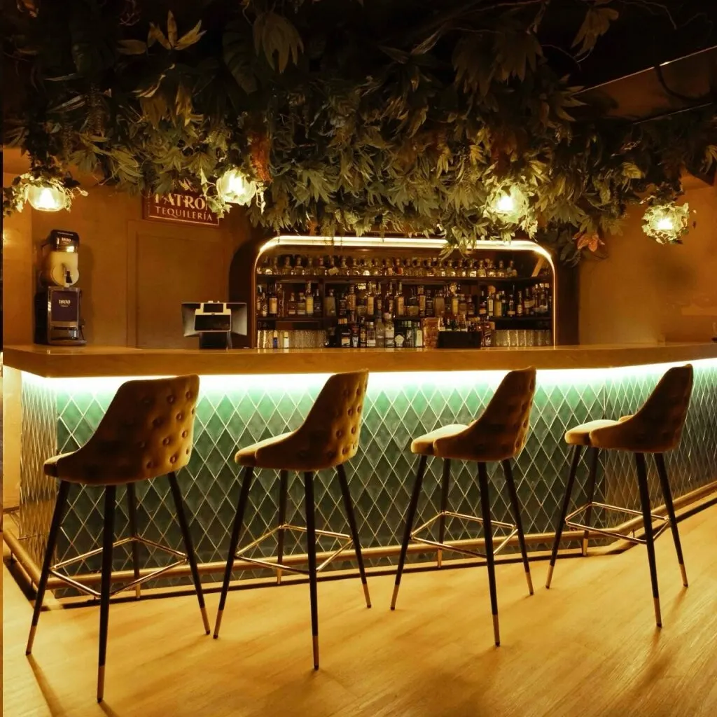 Bar con cuatro taburetes altos frente a barra verde iluminada con patrón de rombos. Botellas al fondo, plantas colgantes y luces cálidas.