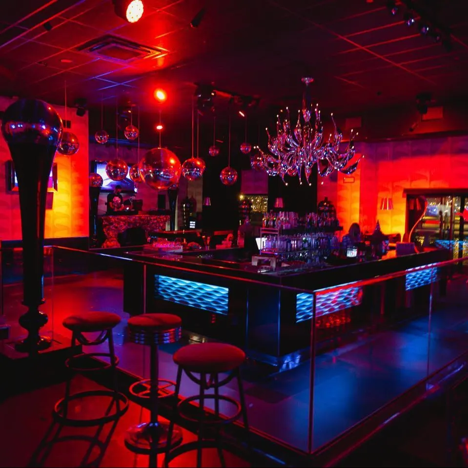 Dux: Este bar vibrante presenta una atmósfera oscura iluminada con luces rojas y azules, decorado con bolas de espejos y un candelabro llamativo sobre una moderna barra central.