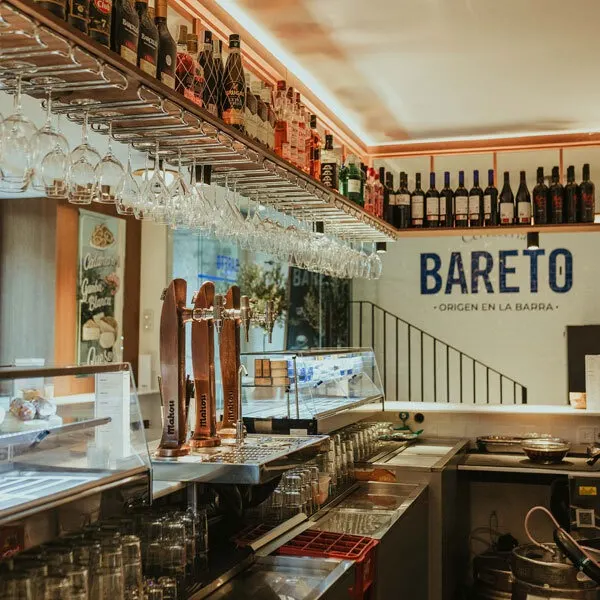 Barra de Bareto vista desde dentro, con grifos de cerveza, copas colgantes y botellas alineadas, bajo el lema “Origen en la barra” al fondo.