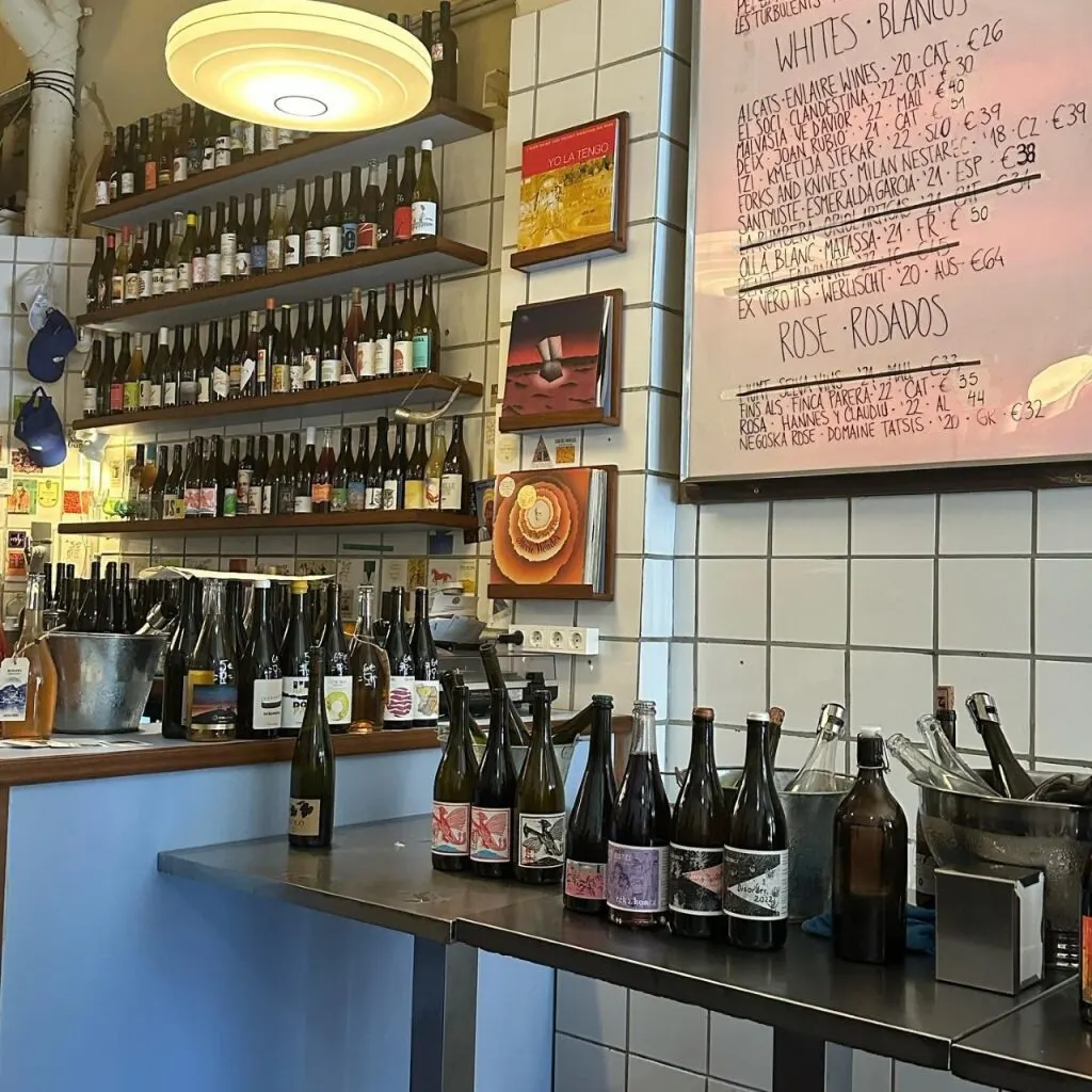 Interior de vinoteca con estanterías de botellas, mesa con vinos alineados, cubitera y cartel con menú de vinos en pared de azulejos.