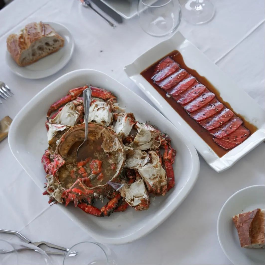 Plato de centollo cocido con caparazón abierto acompañado de sashimi de atún rojo en salsa de soja, sobre mantel blanco y con pan de masa madre.
