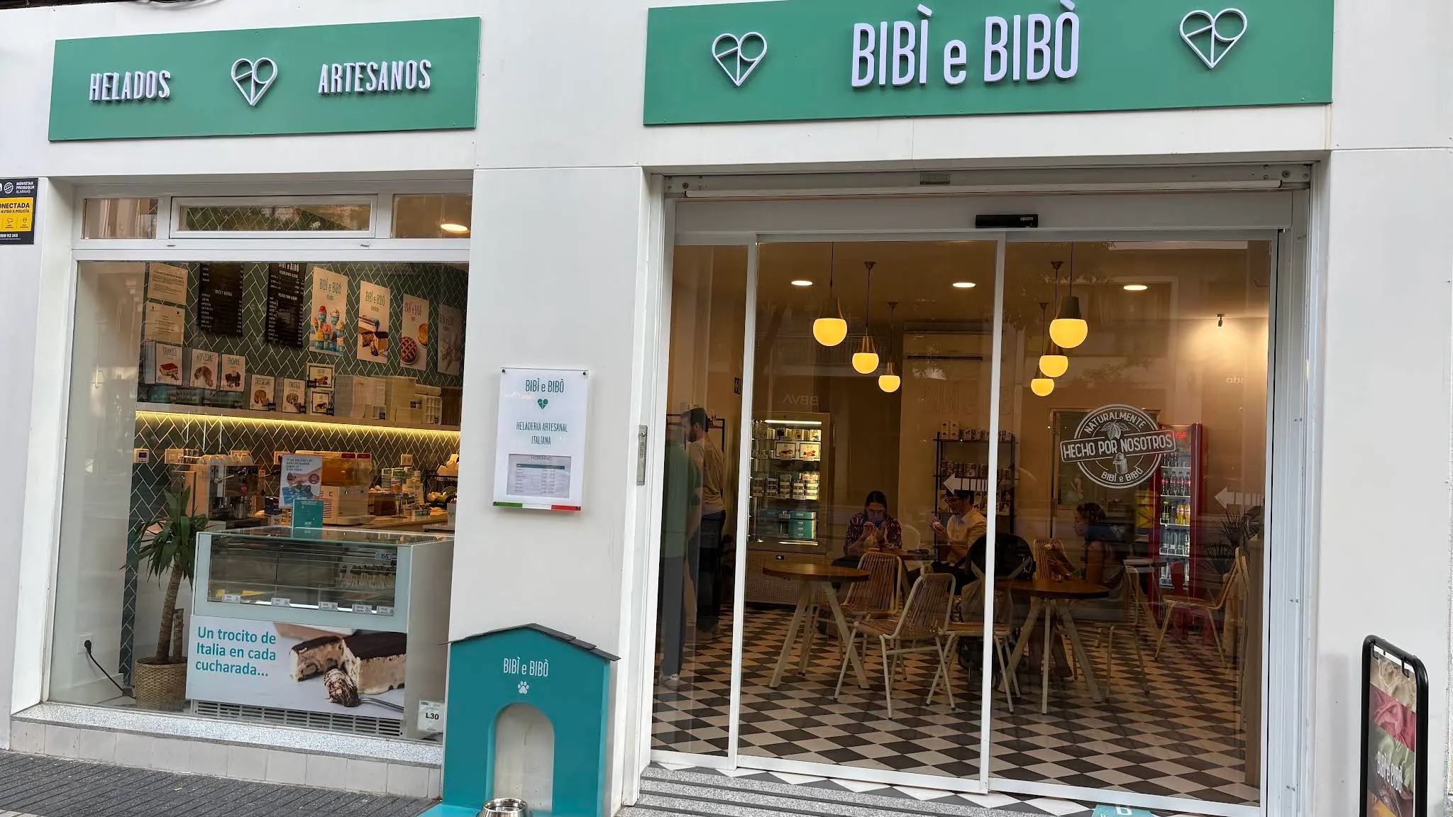Fachada blanca con rótulos verdes, grandes ventanales y acceso acristalado que dejan ver el interior cálido y familiar de Bibi e Bibò Alcalá.