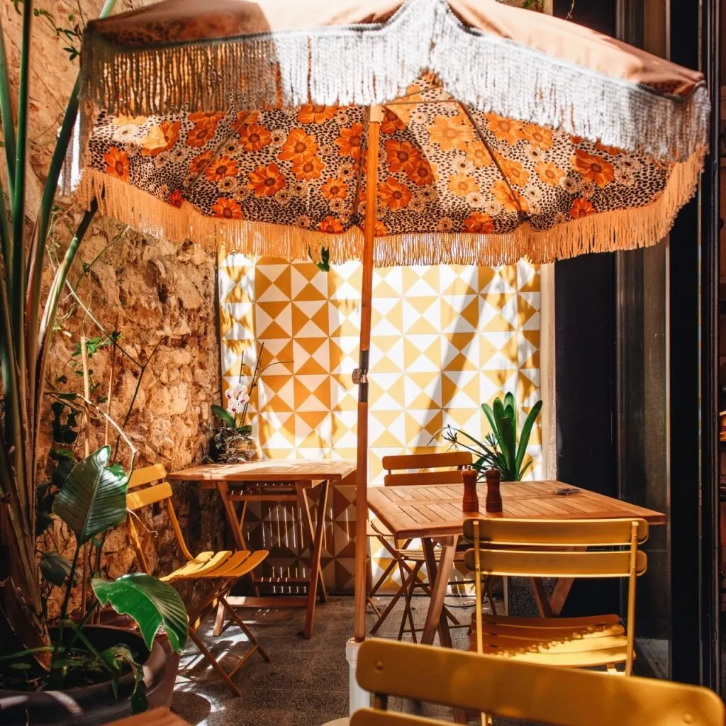 Zona exterior con mesas de madera, sillas amarillas, sombrilla floral con flecos, pared geométrica amarilla y plantas en macetas.