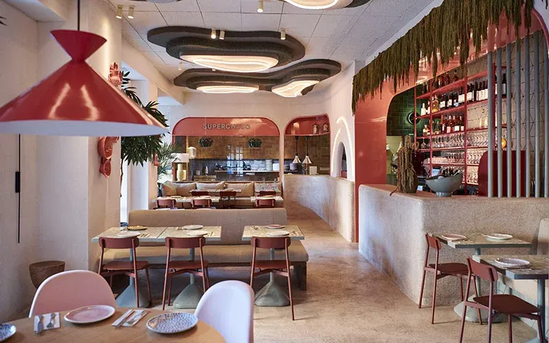 Interior de restaurante con decoración colorida, mesas preparadas, techos ondulados con luces LED y barra con botellas y vegetación colgante.