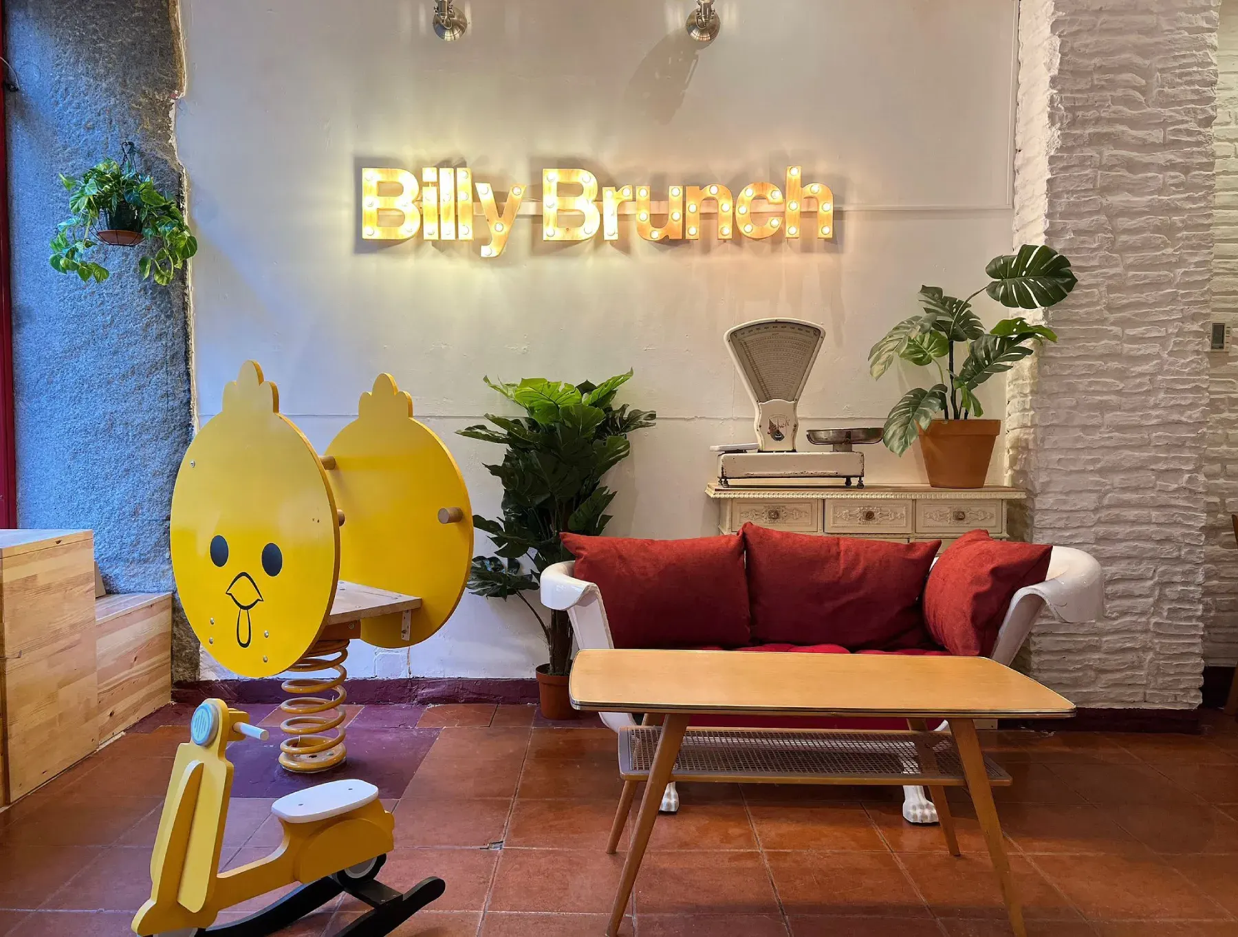 Interior de Billy Brunch con sofá rojo, mesa de madera, balanza antigua, plantas decorativas, juegos infantiles y letrero iluminado en la pared.