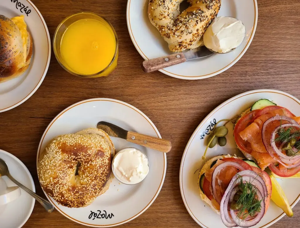 Platos con bagels variados sobre mesa de madera: uno con salmón y verduras, otro con semillas y crema, junto a un vaso de zumo de naranja natural.