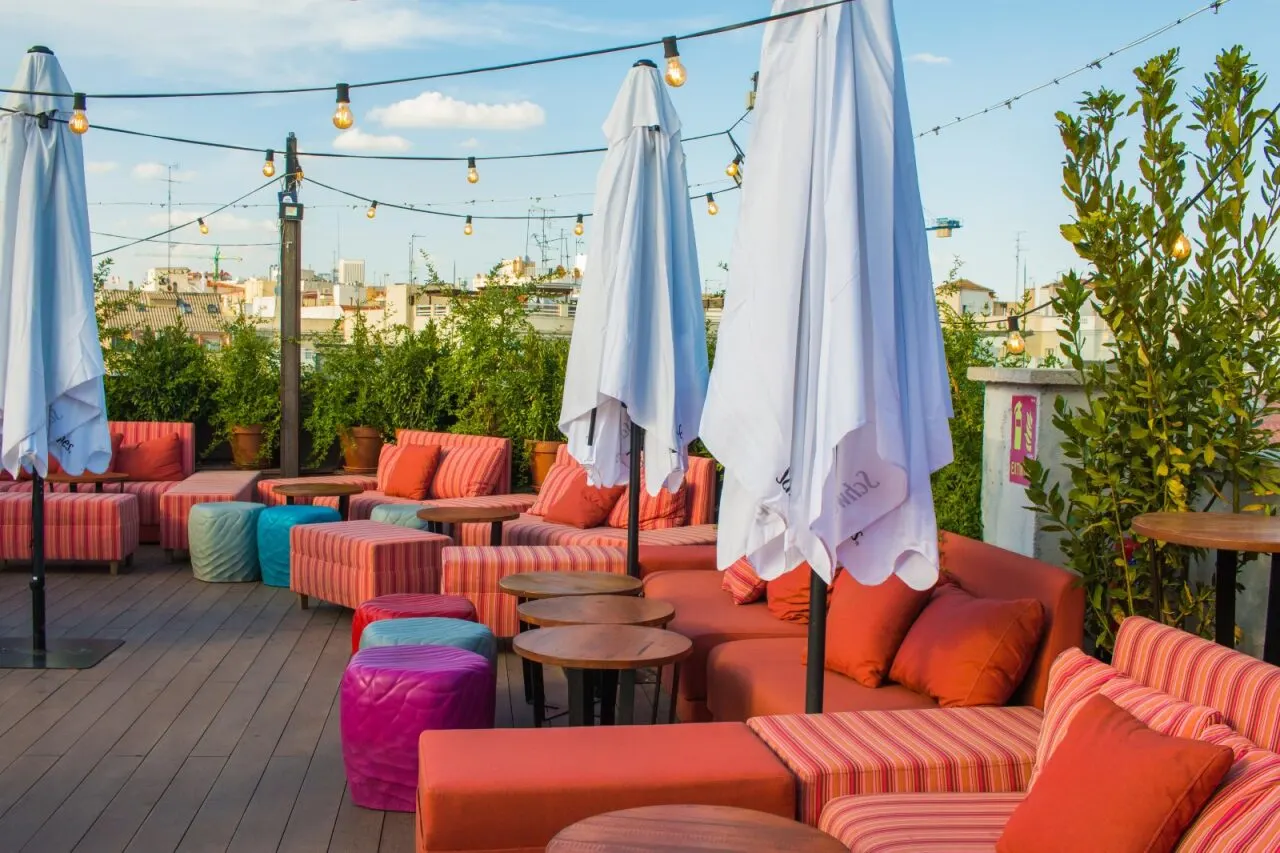 Terraza de Irreverente con sofás naranjas y rayados, pufs de colores y mesas de madera, rodeada de plantas y bombillas colgantes bajo el cielo de Madrid.