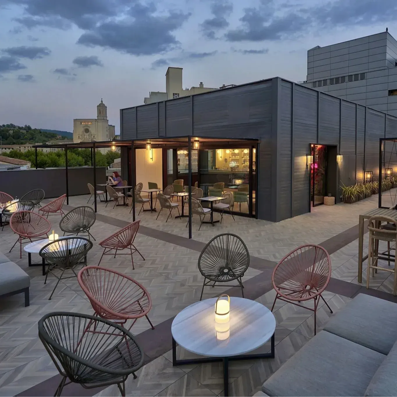 Amplia terraza urbana al atardecer con mesas, sofás y sillas modernas; bar iluminado en un edificio contemporáneo.