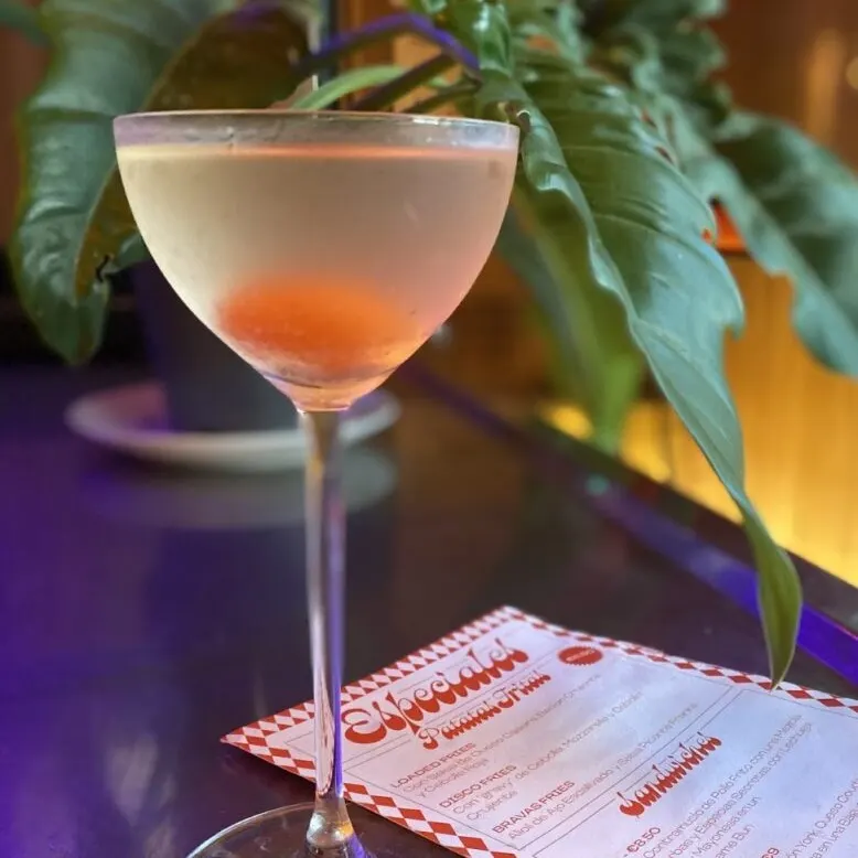 Cóctel claro con rodaja de naranja sobre superficie reflectante, fondo con planta verde de hojas grandes y menús de bebidas parcialmente visibles.