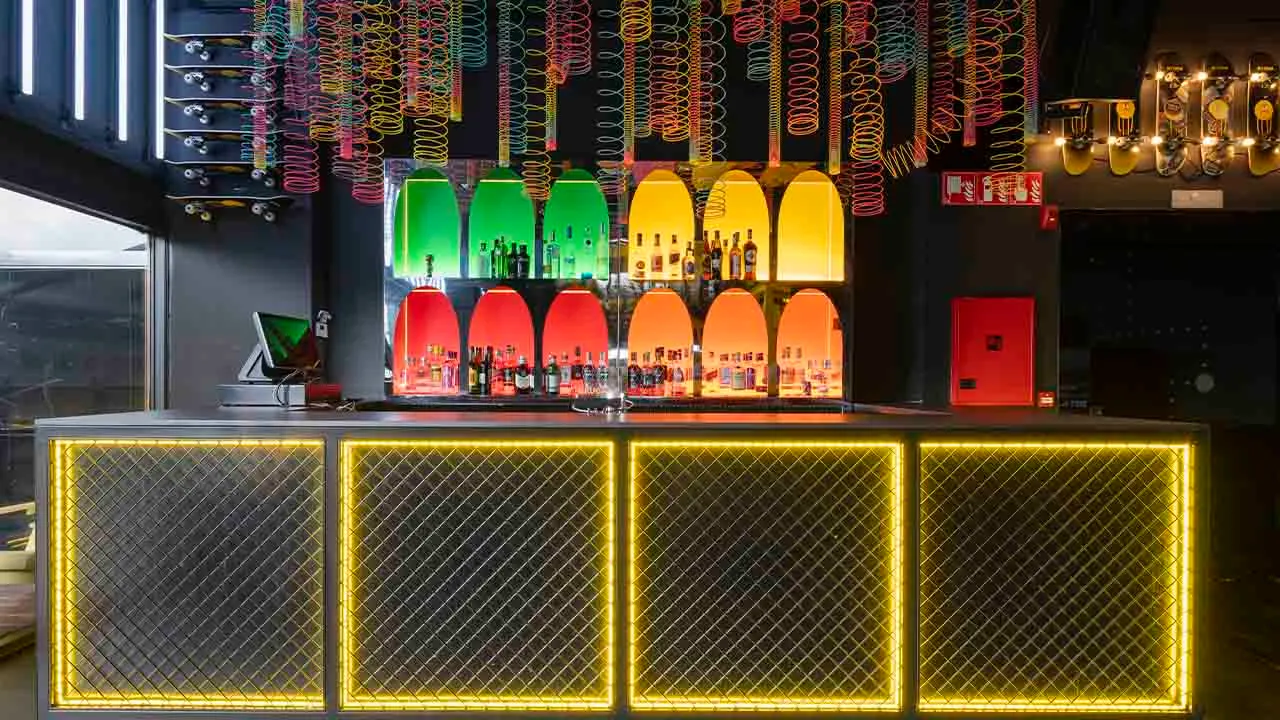 La imagen muestra el interior de HYP3, un espacio moderno y creativo con una estética urbana. Con tablas de skate colgadas, luces de neón y una gran carita sonriente verde en el techo, el ambiente es vibrante y juvenil.