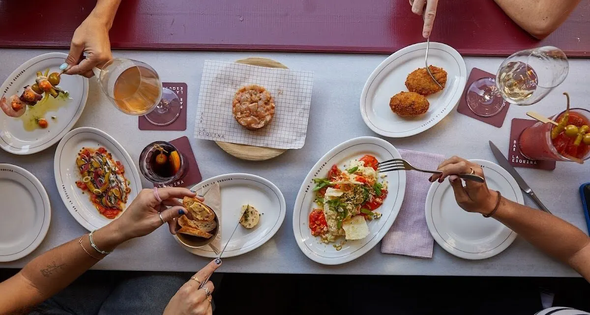 Mesa vista desde arriba con platos variados: brochetas, ensalada, croquetas, tartar y bebidas. Cuatro personas interactúan con la comida.