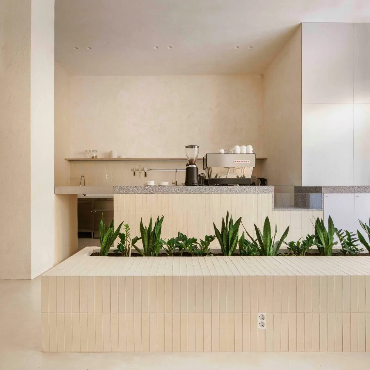 Barra de café minimalista con paredes claras, mostrador de azulejos, cafetera y utensilios. Jardín interior con plantas verdes frente al mostrador.
