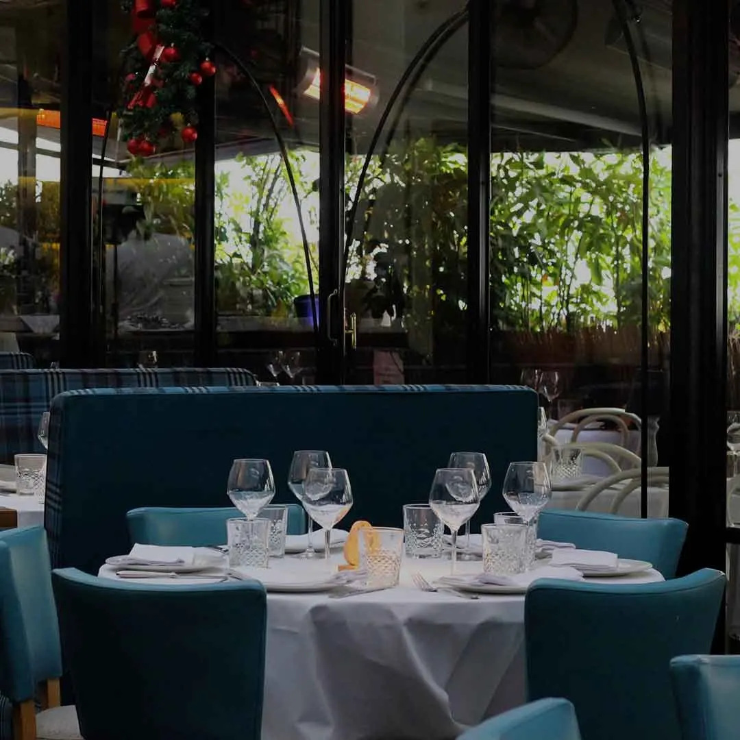 La foto muestra una mesa de comedor interior de la Taberna Los Gallos. Hay mesas con sillas tapizadas en azul y manteles blancos, con copas de vino, servilletas y cubiertos dispuestos.