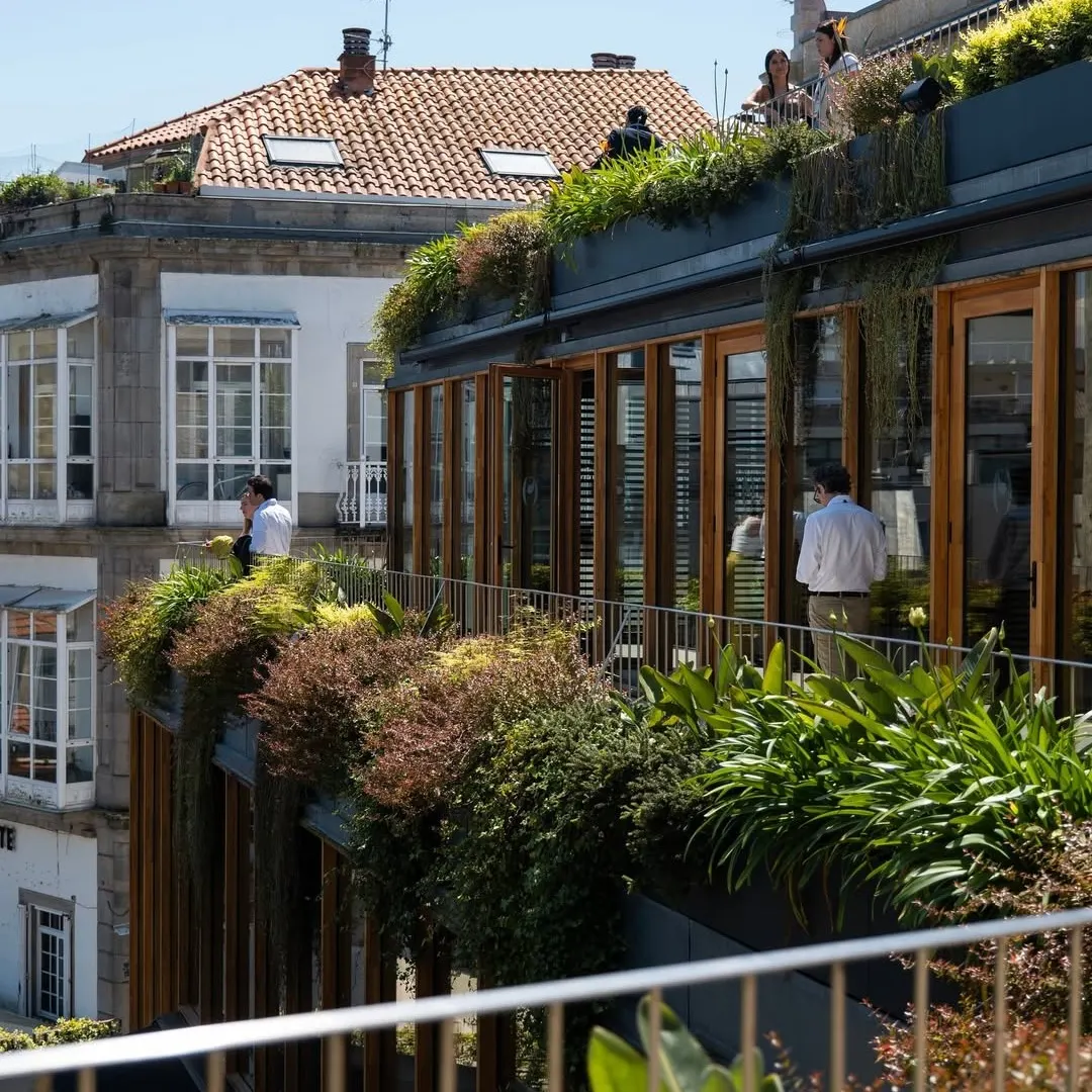 Detalle de la fachada con ventanales de madera y jardineras colgantes, con varias personas paseando por la terraza superior.
