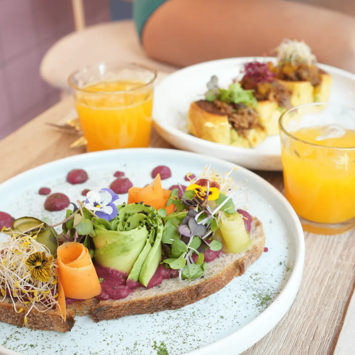Mesa con dos platos: uno con pan, verduras, brotes y flores comestibles; otro con tostada, verdes y carne; dos zumos de naranja.