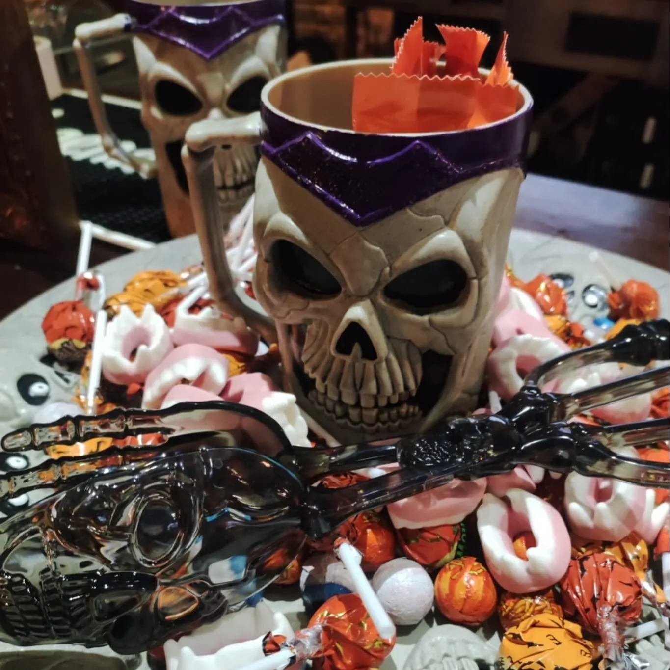 Bandeja con chucherías de Halloween, calaveras, colmillos y caramelos. En el centro, una taza de calavera con envoltorios naranjas.