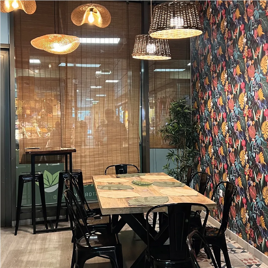 Rincón de La Mala Brunch con mesa de madera, sillas negras, papel tapiz floral y lámparas colgantes que crean un ambiente acogedor.