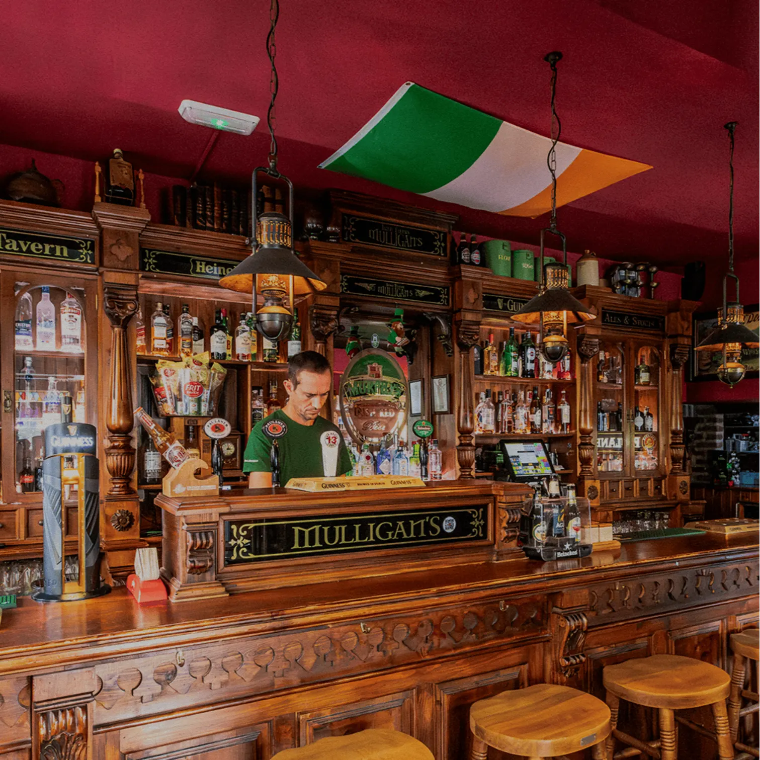 Barra de madera con taburetes altos, botellas alineadas al fondo y camarero sirviendo. Bandera irlandesa colgando del techo.