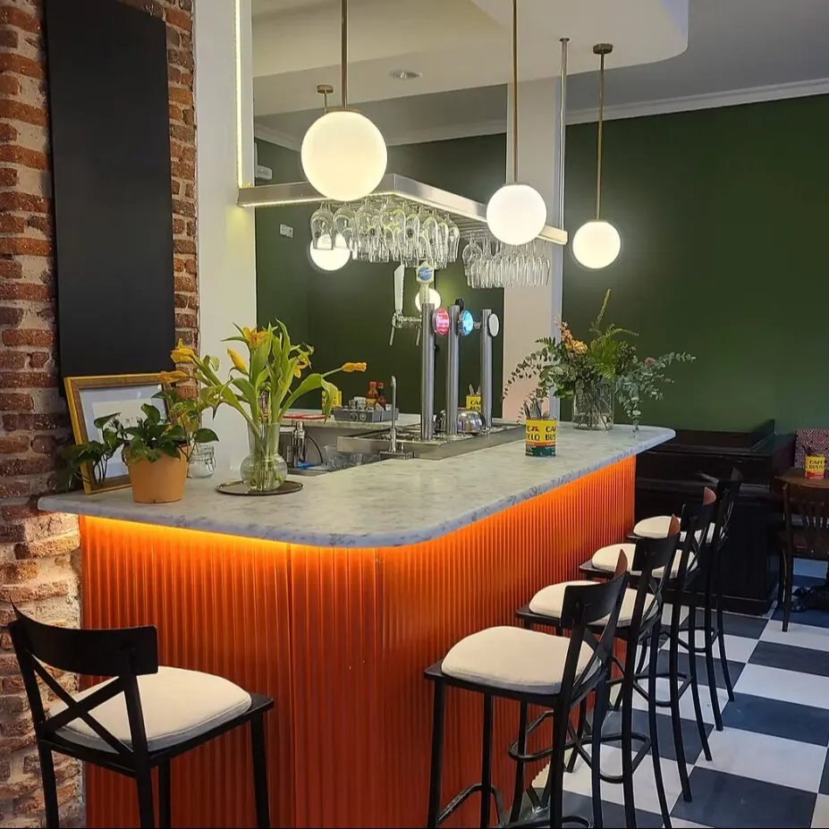 Bar moderno con encimera de mármol, panel iluminado naranja, sillas altas blancas, plantas decorativas y suelo ajedrezado.