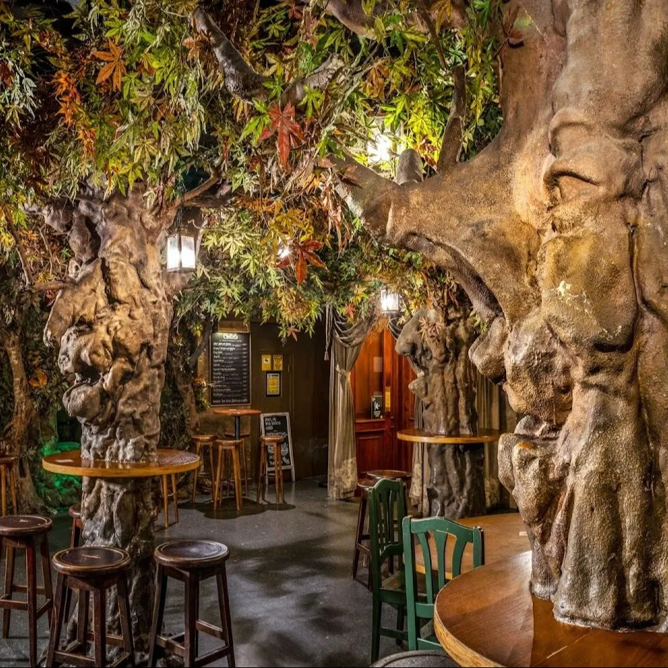 Espacio interior con árboles artificiales, mesas de madera y faroles colgantes; ambiente acogedor tipo café temático.