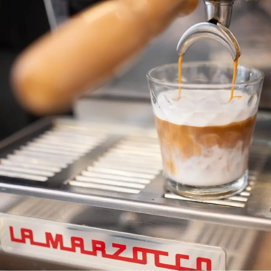 Café con leche vertiéndose en un vaso de cristal sobre una máquina La Marzocco, mostrando el contraste entre el espresso y la leche espumada.