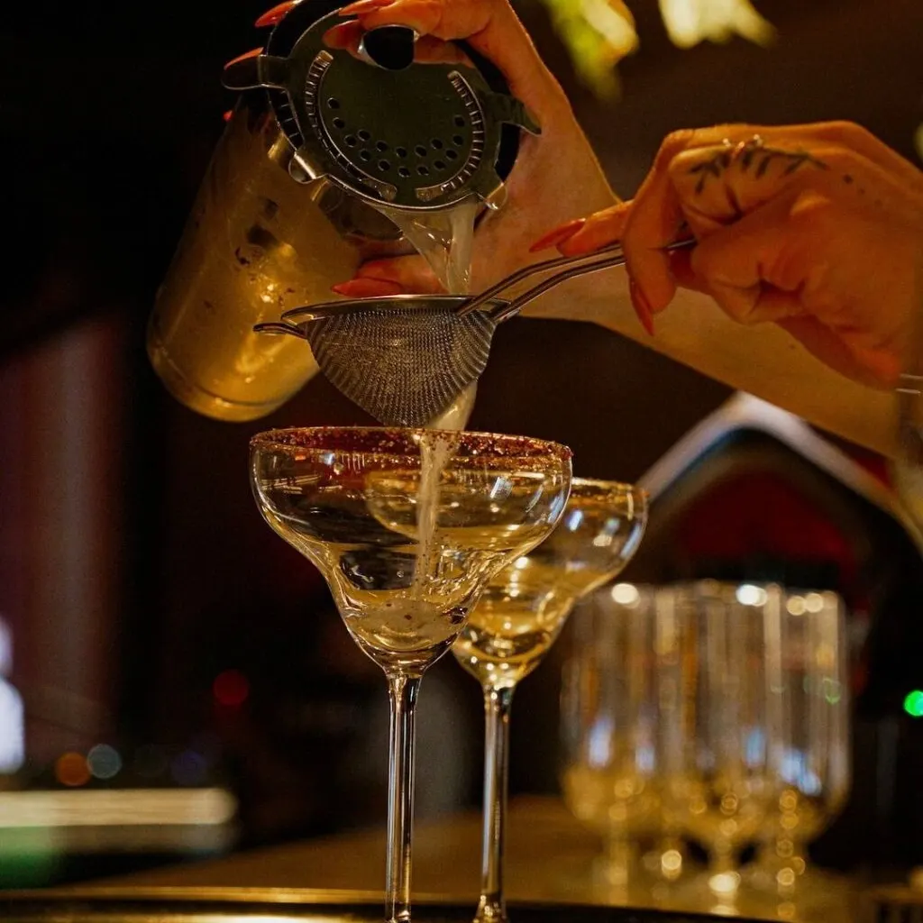 Bartender sirve cóctel filtrado en copa margarita usando colador y coctelera; fondo con más copas en ambiente tenue de bar.