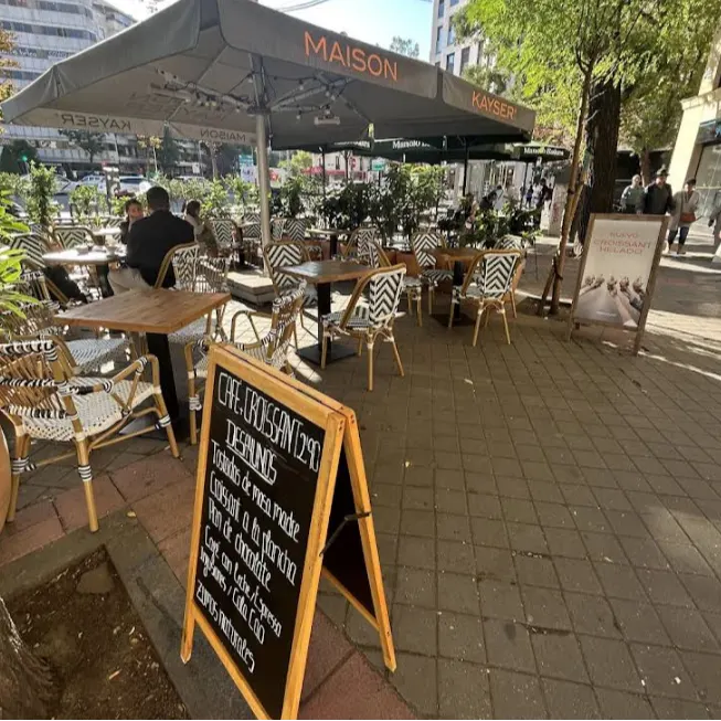 La terraza de Maison Kayser Quevedo tiene sillas de mimbre, mesas de madera y pizarras con desayunos bajo sombrillas.