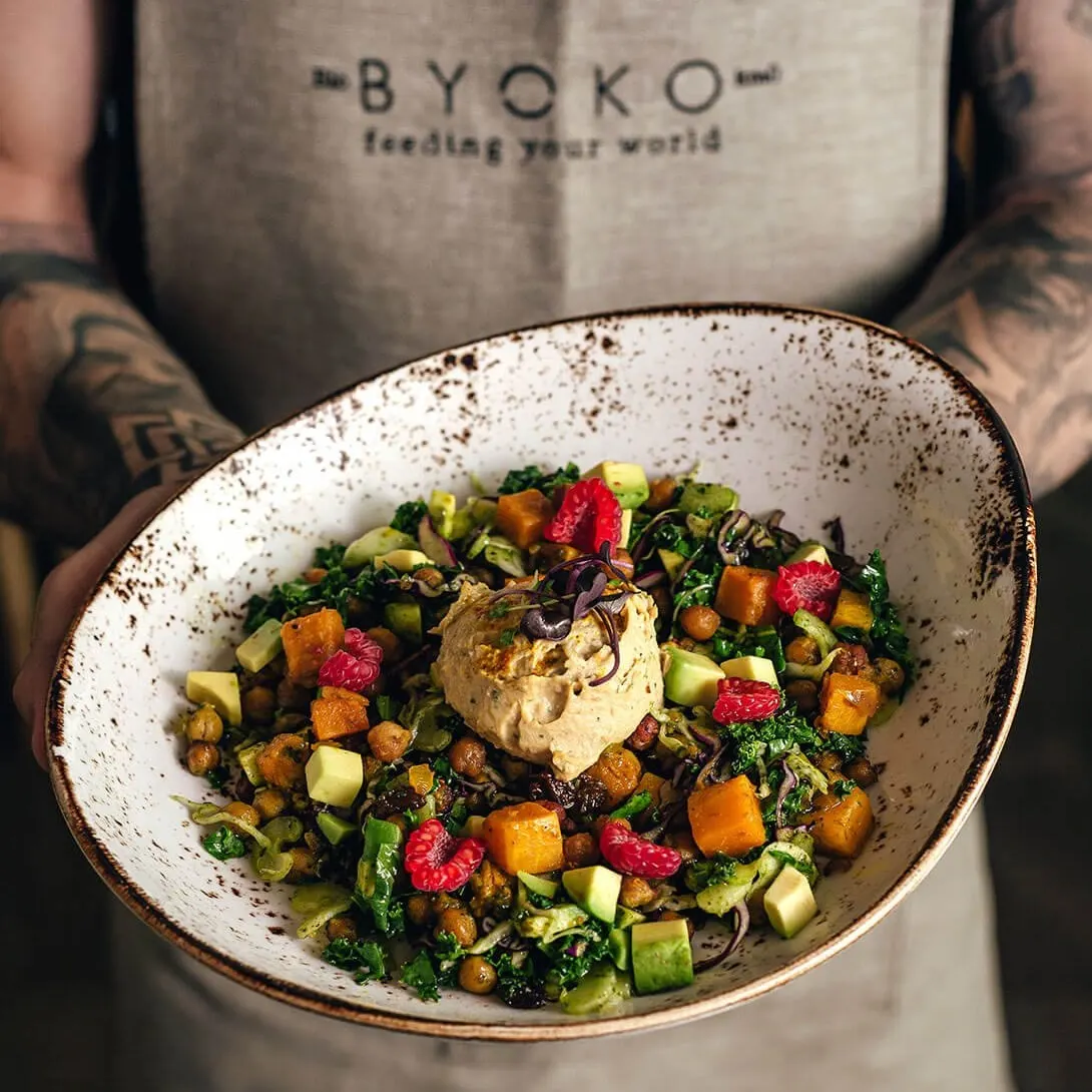 Plato de ensalada colorida con hummus en el centro, sostenido por persona con delantal beige en BYOKO, ideal para una experiencia saludable y visualmente atractiva.