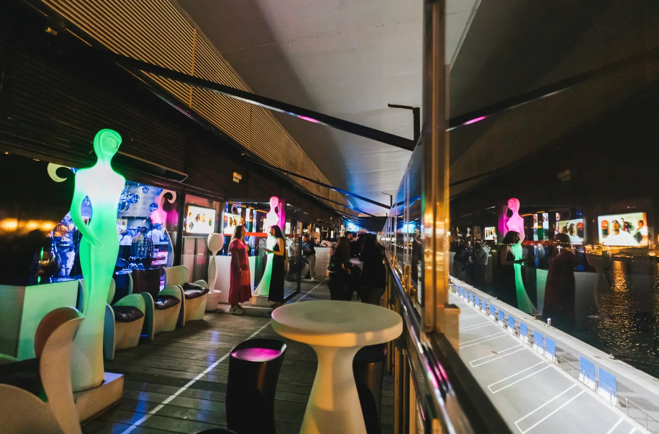 La imagen muestra la terraza del Dux, un espacio moderno y elegante junto al agua. Con figuras iluminadas en colores vibrantes y mobiliario minimalista, el lugar ofrece un ambiente futurista y exclusivo