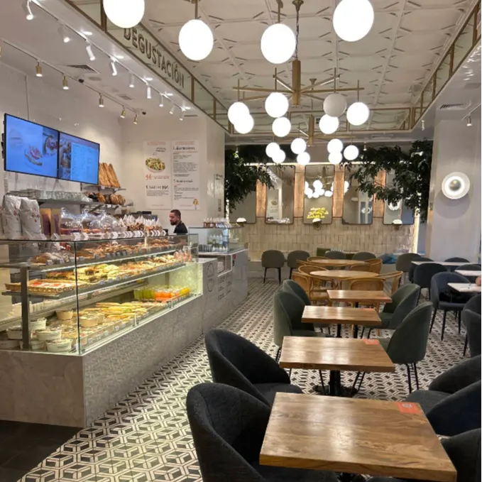 Zona de degustación en Maison Kayser Gaztambide, con vitrinas repletas de bollería francesa, techos altos y lámparas esféricas que iluminan un espacio acogedor.