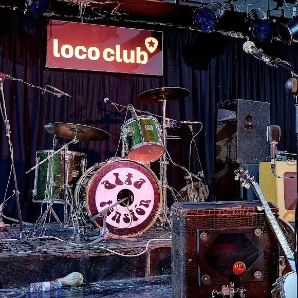Loco Club con escenario listo para vibrantes conciertos en vivo, con batería, guitarras y micrófonos en un ambiente auténtico y underground.