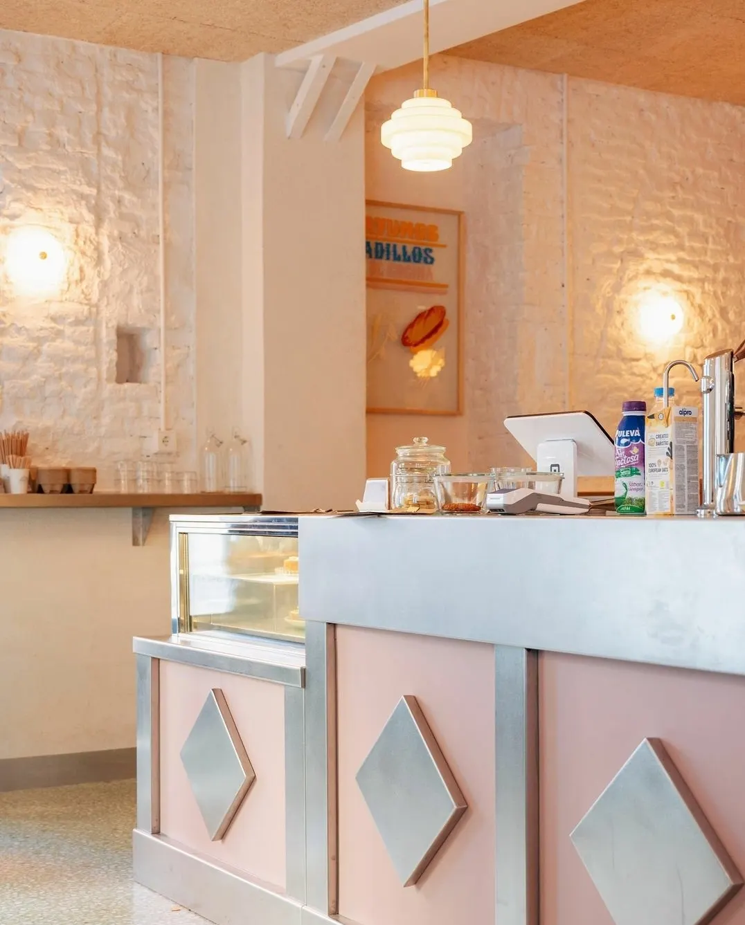 Mostrador retro en tonos rosa y acero con detalles geométricos, vitrina con dulces, paredes de ladrillo blanco y lámpara colgante en Trava de Noche.