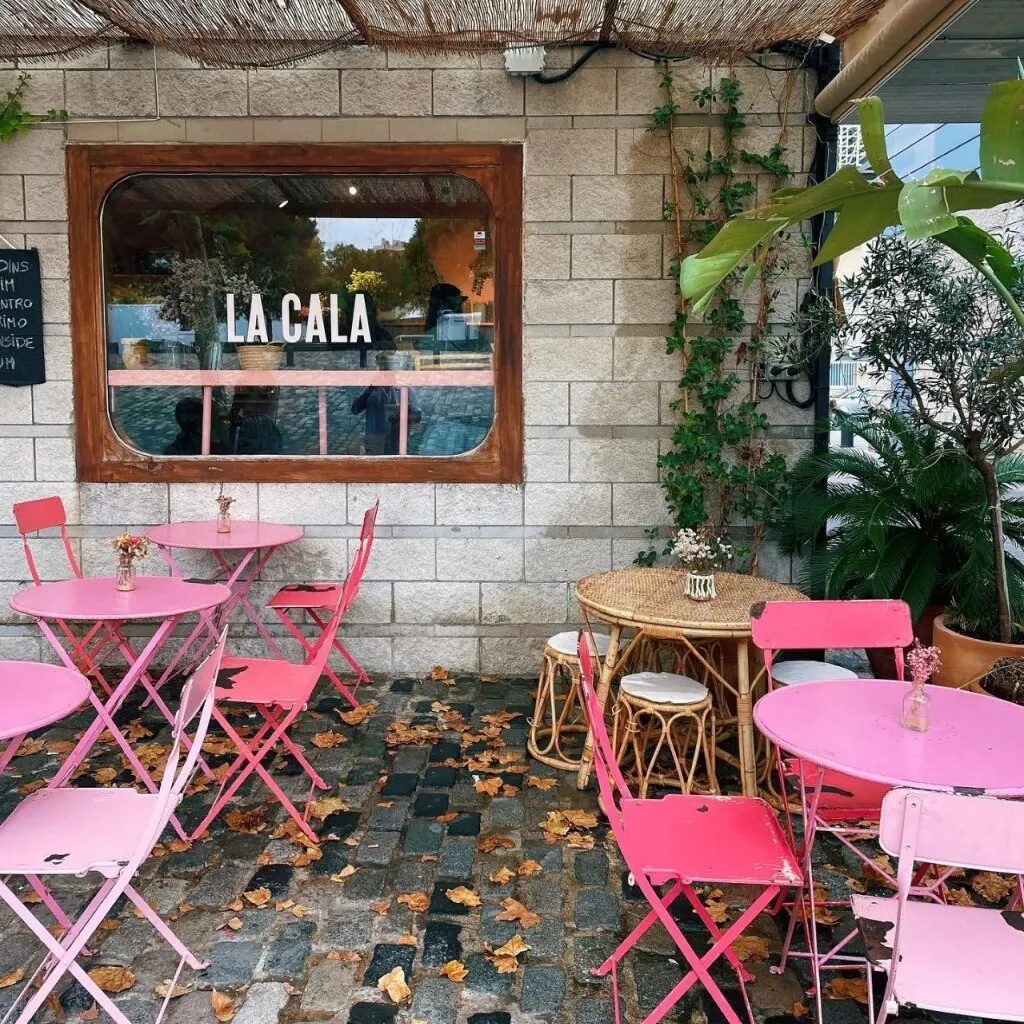 Terraza de LA CALA con mesas rosas, sillas metálicas, hojas caídas, muebles de mimbre y plantas sobre suelo empedrado.