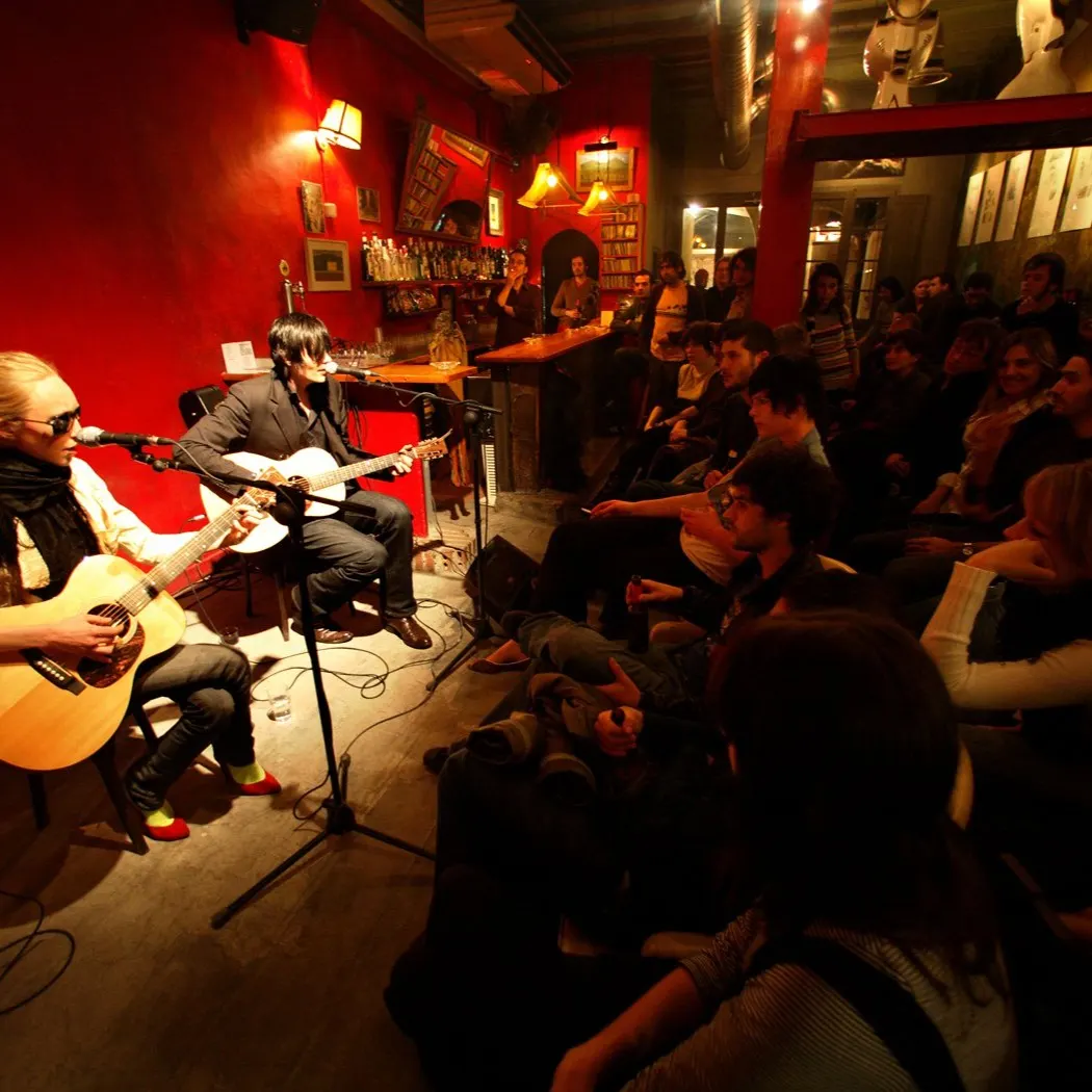 La imagen muestra a dos músicos tocando guitarras acústicas en un ambiente íntimo, rodeados de un público atento en un local con paredes rojas y una atmósfera cálida.