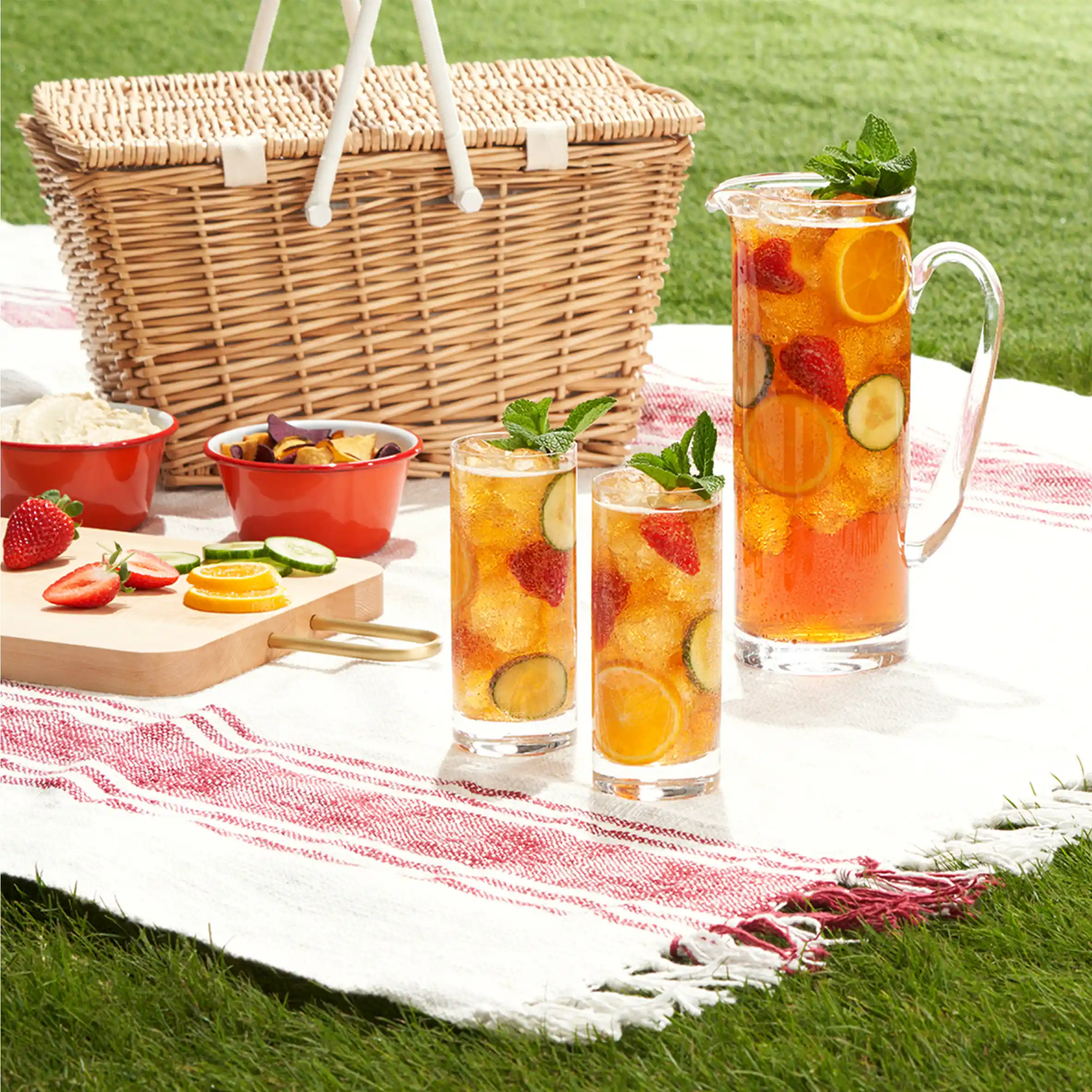 Picnic de verano con una cesta de mimbre, una jarra y vasos llenos de una bebida refrescante con frutas y menta, sobre un mantel a cuadros rojos y blancos